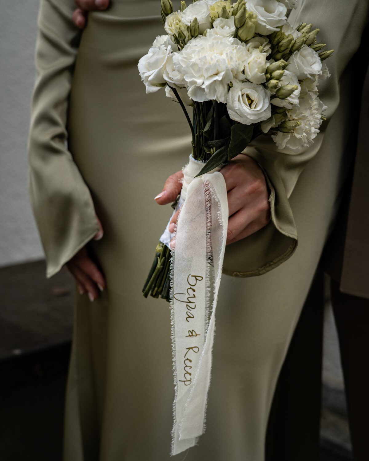 Braut im salbeigrünen Kleid hält weißen Brautstrauß mit beschriftetem Band